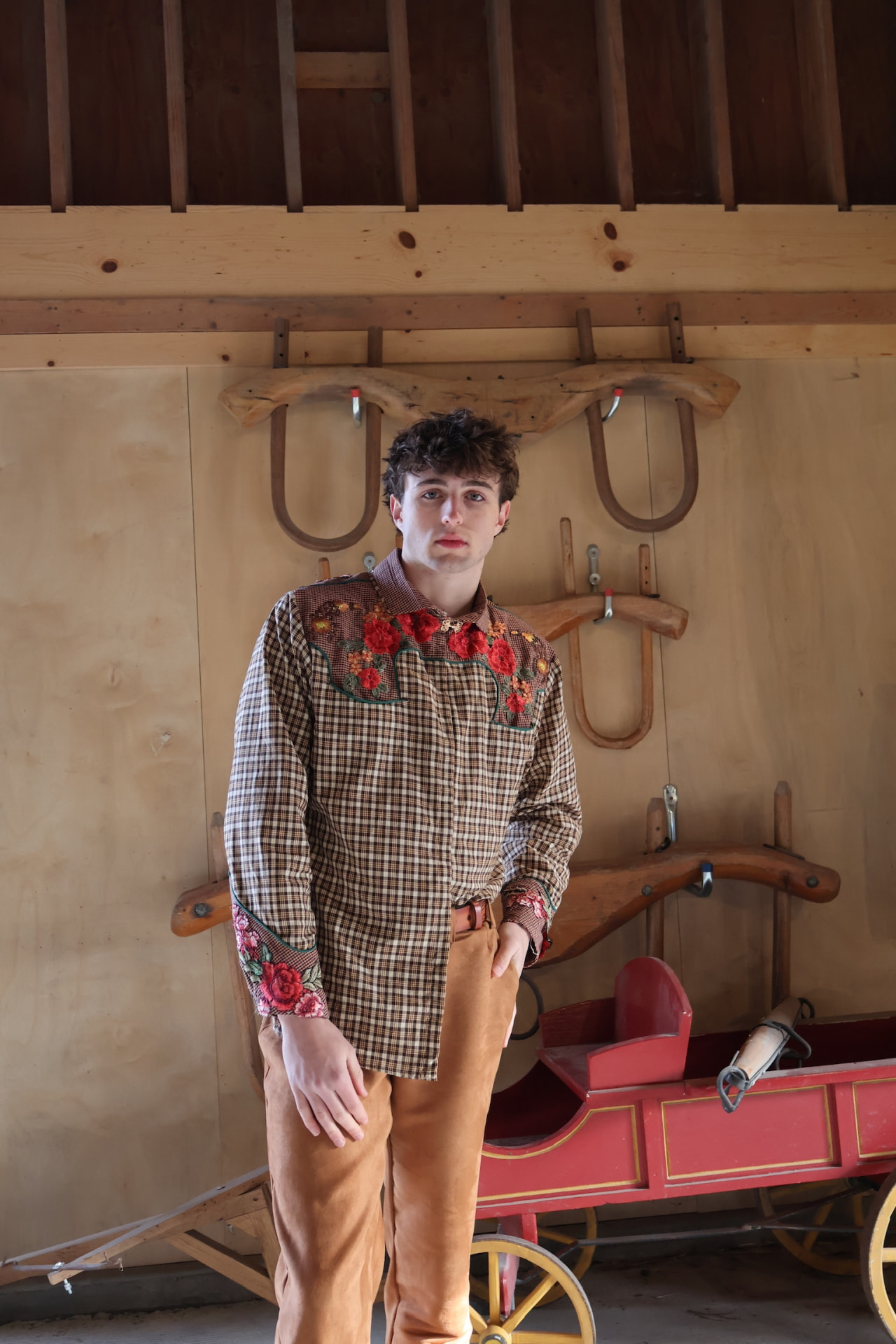 Model in an embroidered plaid shirt and tan trousers in a rustic wooden interior